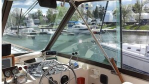 Interior de un barco con timón, paneles de control y vistas a un puerto deportivo a través de amplias ventanas.