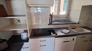 Ett kompakt kök med vita skåp, en svart bänkskiva, en vattenkokare, ett handfat och ett fönster med utsikt över omgivningen.