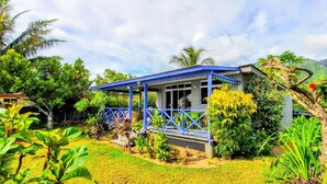 A house with a blue roof and a covered porch, surrounded by lush greenery and a mountainous backdrop.