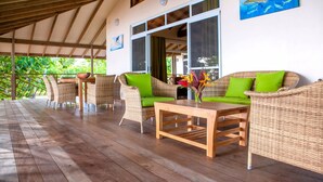 A covered outdoor seating area with wicker furniture, a wooden table, and large windows. - Tahaa - Villa Tehere Dream Beach Front (Taha'a)