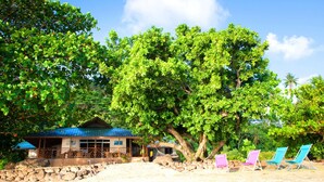 A beach house with a blue roof, surrounded by lush green trees and a sandy beach. There are colorful beach chairs and a stone wall in the foreground. - Tahaa - Villa Tehere Dream Beach Front (Taha'a)