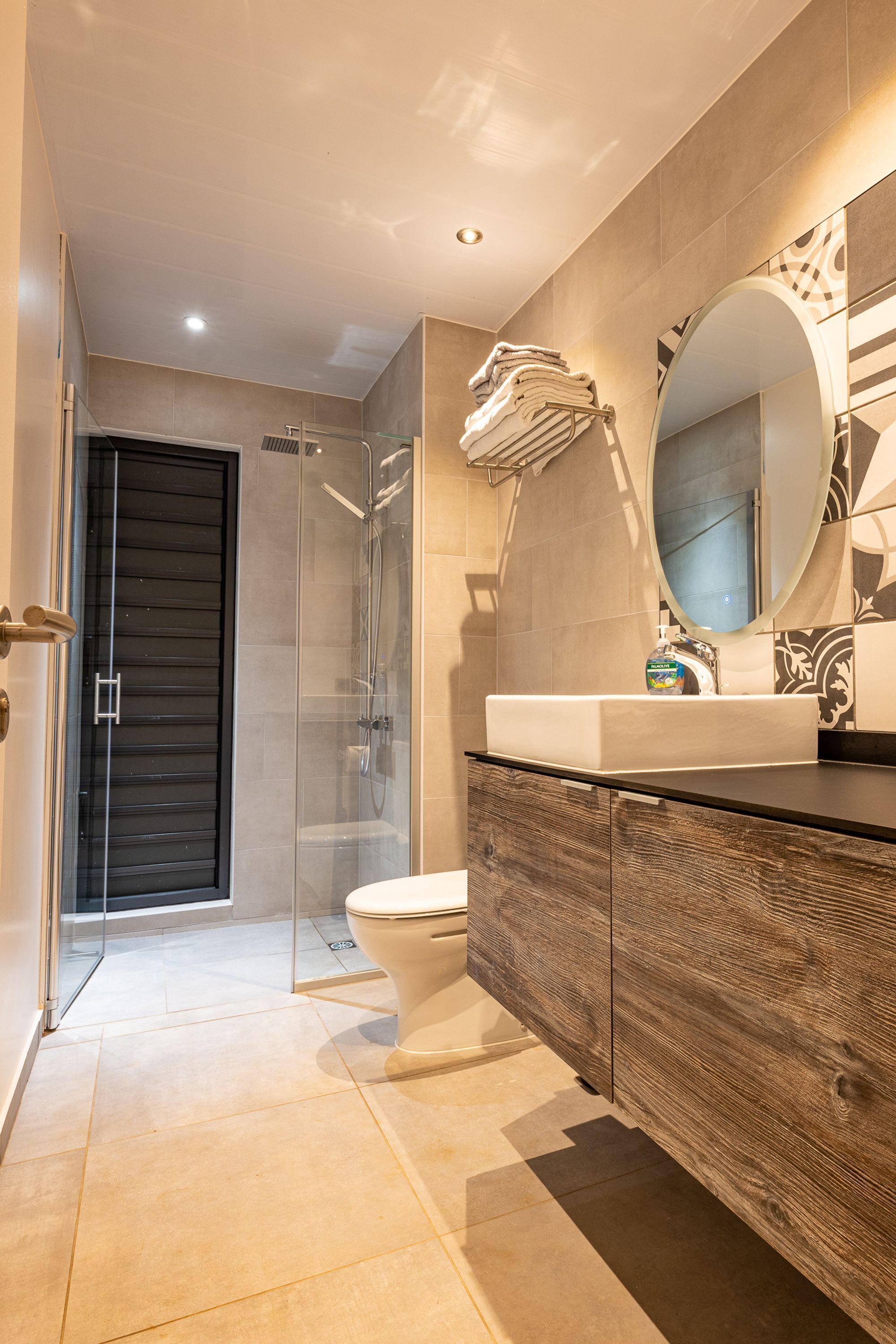 Une salle de bain moderne avec une douche à parois de verre, des toilettes blanches et un meuble-lavabo en bois doté d’un grand miroir.