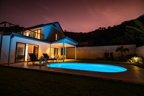 Een modern huis met een zwembad, buiten zitplaatsen en een goed verlicht terras bij zonsondergang.