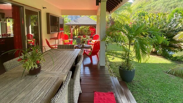Un espace de restauration en plein air avec une table en bois et des chaises en osier, entouré d’une végétation luxuriante et offrant une vue sur une colline.