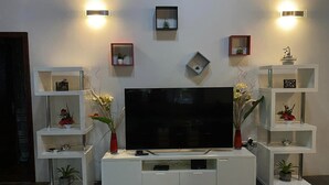 Un salon moderne doté d’un grand téléviseur à écran plat placé au centre d’un meuble de rangement blanc, flanqué de deux étagères blanches ornées d’objets décoratifs et de petites plantes en pot.