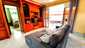 A living room with a grey sofa, wooden entertainment unit, and a dining area visible through a glass door. - Tahiti - Lihei Pool House (Faaa)