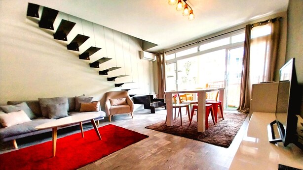 A modern living room with a sofa, a coffee table, a red rug, a dining area with a table and chairs, and a staircase with black railings. - Tahiti - Urban Escape (Papeete)