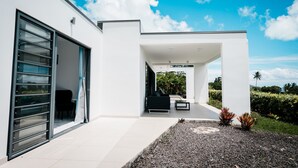 Una casa moderna con un amplio ventanal corredera, un patio con sofá negro y mesa de centro, y vistas a exuberante vegetación.