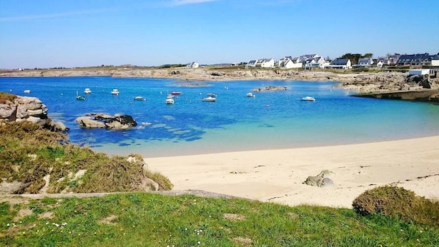 Une plage avec une eau d’un bleu limpide, des bateaux et des maisons en arrière-plan.