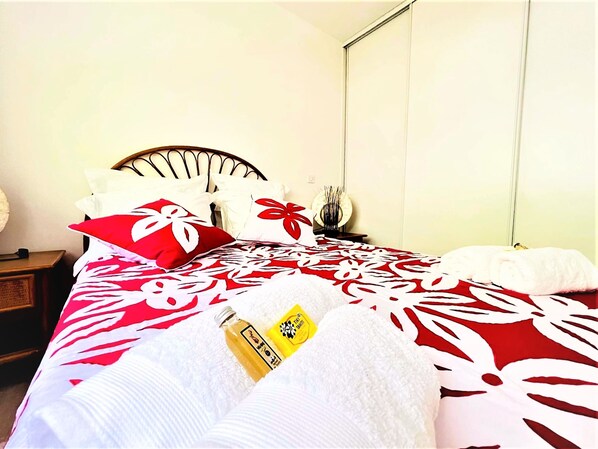 A neatly made bed with a red and white floral bedspread, a bottle of lotion, and a white towel. - Tahiti - Toke Condo in Faa a (Faaa)