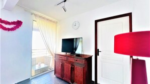 A room with a TV on a wooden cabinet, a red lamp, a white door, and a clock on the wall. - Tahiti - Toke Condo in Faa a (Faaa)