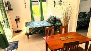 Ein Zimmer mit Holztisch, Stühlen, einem Bett mit Blumenmuster und einem Deckenventilator, sowie einem Fenster mit Vorhängen.