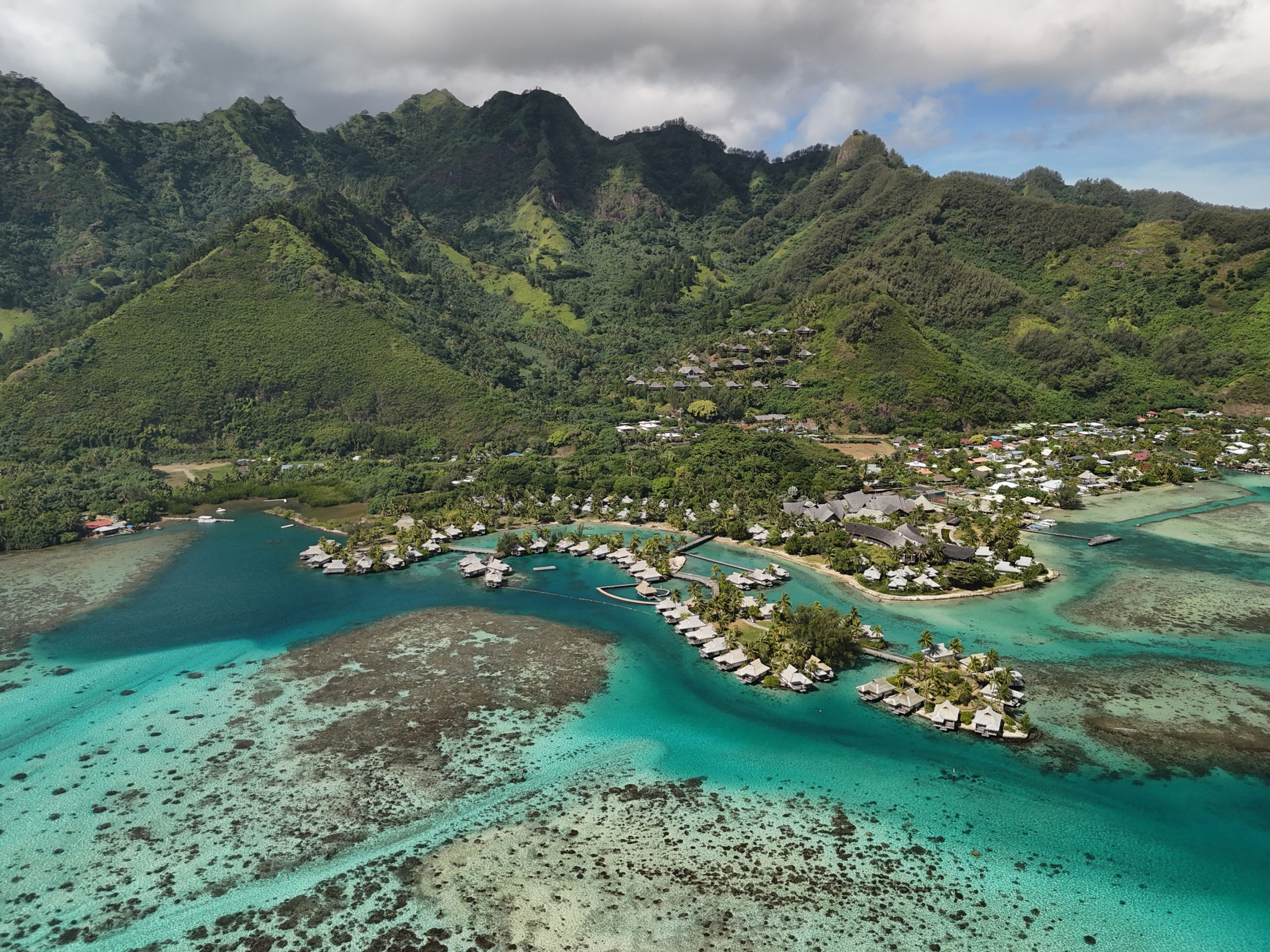 An aerial view of a resort with overwater bungalows, clear turquoise waters, and lush green mountains in the background.