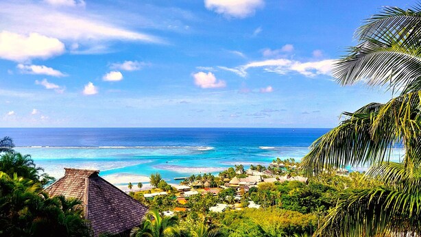 A coastal view with a clear sky, turquoise waters, and a small settlement with traditional thatched-roof buildings. - Moorea - Villa Ana Mua (Moorea-Maiao)