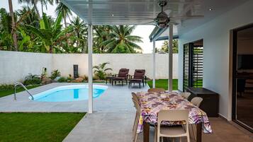 Relax by the pool surrounded by lush greenery and palm trees, with a dining area and ceiling fan for comfort.