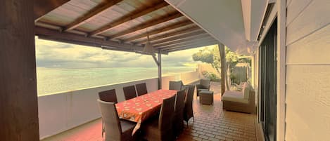 A covered patio with a table and chairs, a view of the sea, and a brick floor.