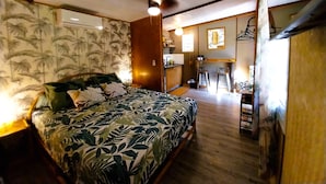 A bedroom with a bed, a wooden ceiling, a wall with a tropical wallpaper, a small kitchenette, and a bathroom visible through an open door. - Moorea - Tropical Nest (Moorea-Maiao)