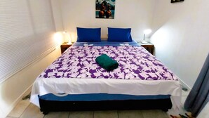 A bedroom with a bed, two blue pillows, a purple floral bedspread, a green pillow, two bedside lamps, an air conditioner, and a wall-mounted picture. - Tahiti - Matavai Bay Cottage (Arue)