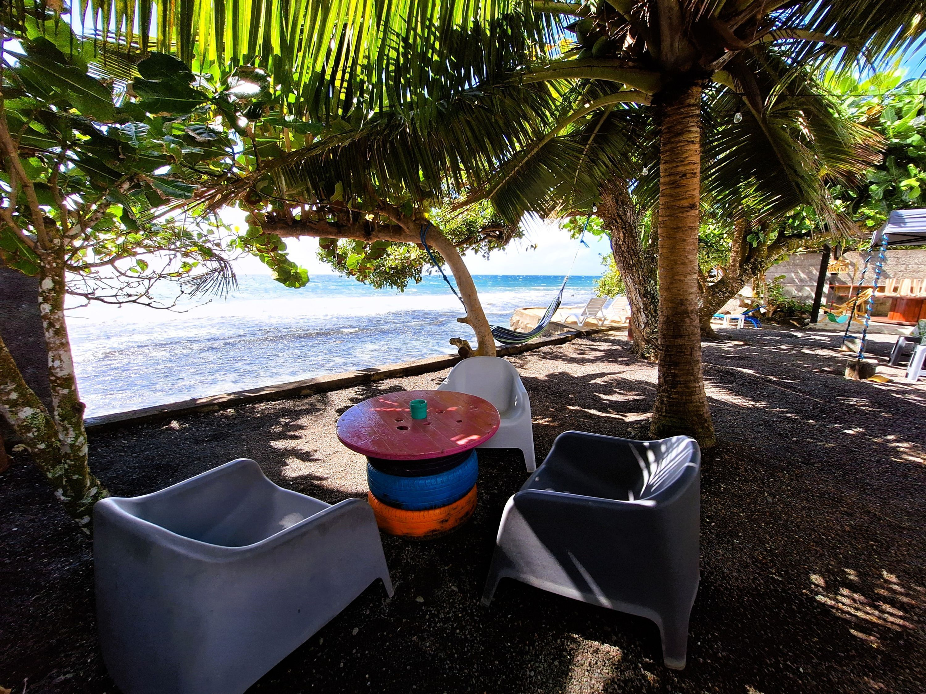 A beachside seating area with two plastic chairs, a round table, and a hammock under a palm tree, overlooking the sea.
