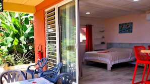 A room with a bed, a table with chairs, and a sliding glass door leading to a balcony with plants. - Tahiti - Terevete Pool Studio (Arue)