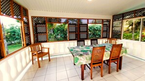 A covered patio area with a dining table and chairs, surrounded by wooden lattice panels and large windows offering views of greenery. - Tahiti - Haumaru Beach Fare (Mahina)