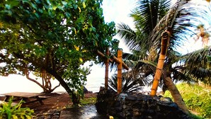 A stone pathway leading to a wooden bench under a tree, with a palm tree and bamboo structure in the background. - Tahiti - Haumaru Beach Fare (Mahina)