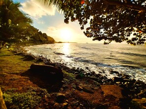 A beach with rocks and vegetation, a clear sky, and the sun setting over the ocean. - Tahiti - Haumaru Beach Fare (Mahina)