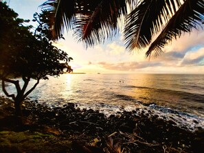 A beach scene with a rocky shoreline, palm trees, and a sunset over the ocean. - Tahiti - Haumaru Beach Fare (Mahina)