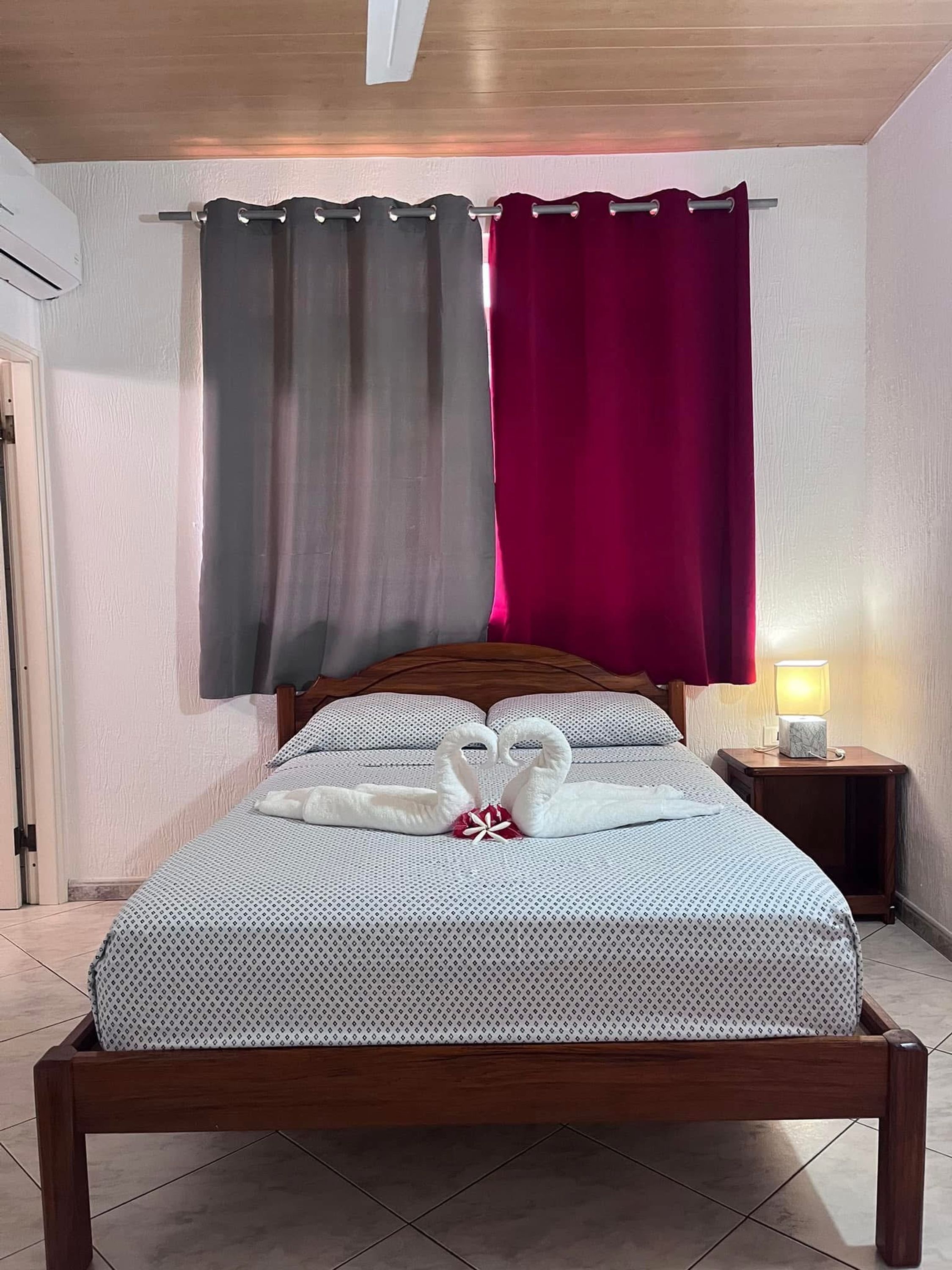 A neatly made bed with a heart-shaped towel arrangement, a wooden headboard, a bedside table with a lamp, and a window with two curtains.