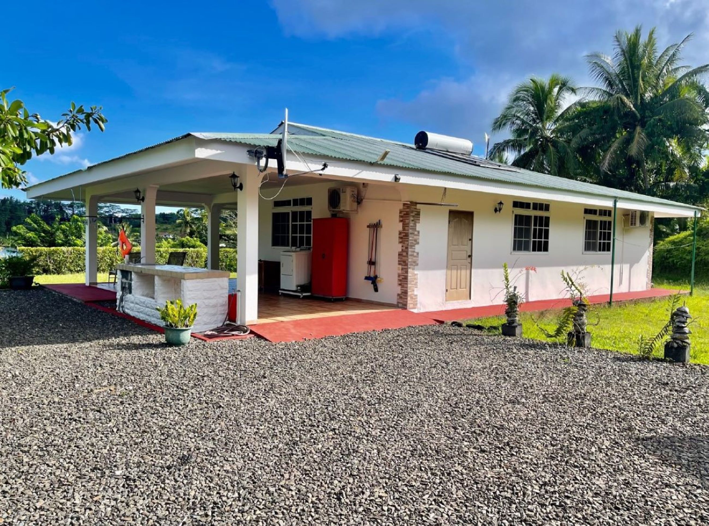 Una construcción de un piso con porche cubierto, puerta roja y un pequeño jardín con grava y plantas en macetas.