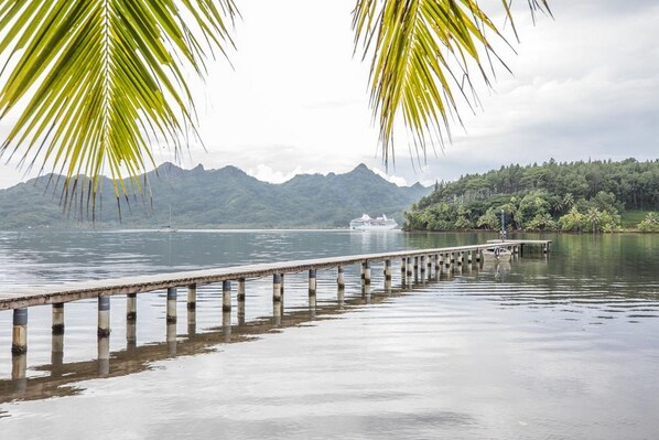 A pier extending into calm water with a boat in the distance, surrounded by lush greenery and mountains.