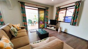 Een woonkamer met een bank, een salontafel en een tv-meubel. Er is een schuifpui die leidt naar een buitenruimte met stoelen en een tafel.