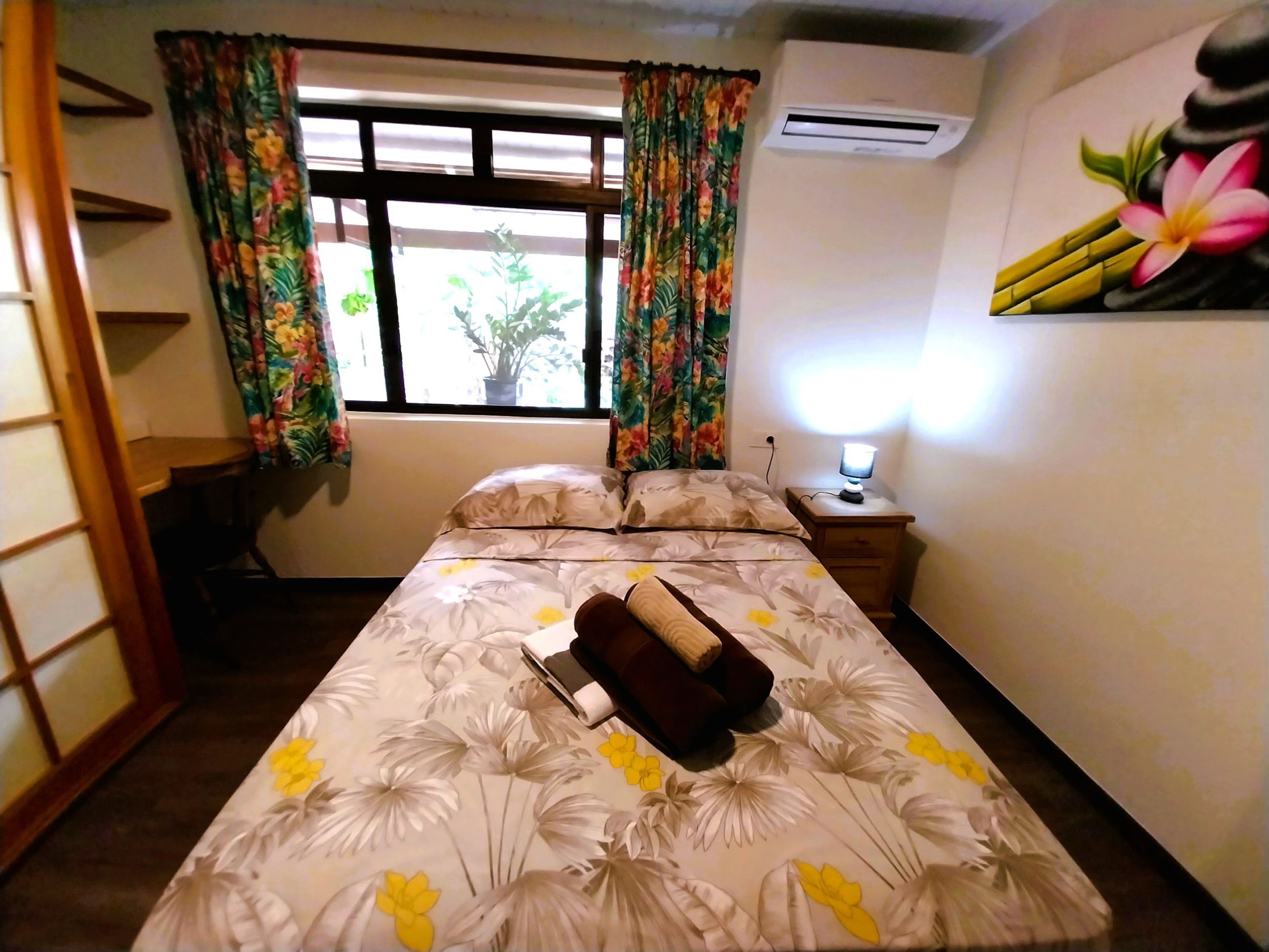 Een slaapkamer met een bed, een nachtkastje met lamp, een raam met gordijnen en een wandairconditioner.
