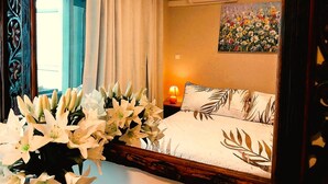 Ein Schlafzimmer mit Bett, Nachttisch, Lampe, einem Fenster mit Vorhängen, einer Wand-Klimaanlage und einem Blumenbild.