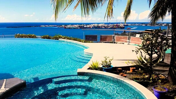 Ein gebogener Infinity-Pool mit klarem, blauem Wasser, umgeben von einer Terrasse mit Pflanzen in Töpfen und Blick auf das Meer und den Hafen.
