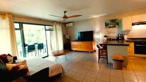 Ein modernes Wohnzimmer mit einer Couch, einem Couchtisch und einem Fernseher. Es gibt einen Balkon mit einer Schiebetür und einem Deckenventilator.