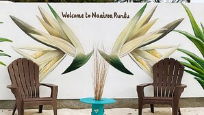 Deux chaises en bois, placées face à face, sont séparées par une table turquoise. L’ensemble est posé sur une surface sablonneuse, avec en arrière-plan un mur orné d’une pancarte de bienvenue et d’illustrations de plantes.