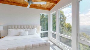 A bedroom with a large bed, a ceiling fan, and large windows offering a view of the outdoors.