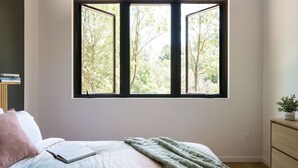 A bedroom with a large window, a bed with a green blanket, a bedside table with a plant, and a dresser. - Tiny Home in the Woods Hot Tub 4 Miles to DT Haus 50 (Asheville)