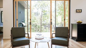 A room with two green upholstered chairs facing a glass table with a tray of wine glasses. A sliding glass door leads to an outdoor area with a view of trees and a barbecue grill. - Tiny Home in the Woods Hot Tub 4 Miles to DT Haus 50 (Asheville)