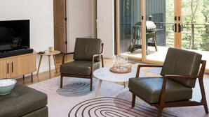 A modern living room with a flat-screen TV, two leather armchairs, a small coffee table, and a large sliding glass door leading to an outdoor area. - Tiny Home in the Woods Hot Tub 4 Miles to DT Haus 50 (Asheville)