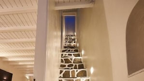 Una escalera con escalones de piedra irregulares que ascienden, paredes blancas y un techo plano con vigas de madera.