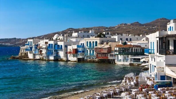 Un pueblo costero con edificios blancos y detalles azules, una playa con mesas y sillas, y un cielo azul despejado.