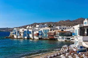 A coastal town with white buildings with blue accents, a beach with tables and chairs, and a clear blue sky. - Blanco Rooms Mykonos (Mykonos)