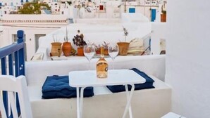 Una terraza en la azotea con mobiliario blanco, una mesa con un florero y una copa de vino, y vistas a un pueblo con casas encaladas.