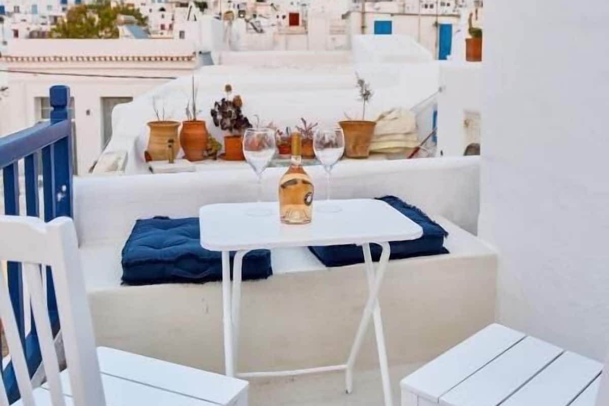 Um terraço no telhado com móveis brancos, uma mesa com um vaso e taça de vinho, e vista de uma cidade caiada de branco.