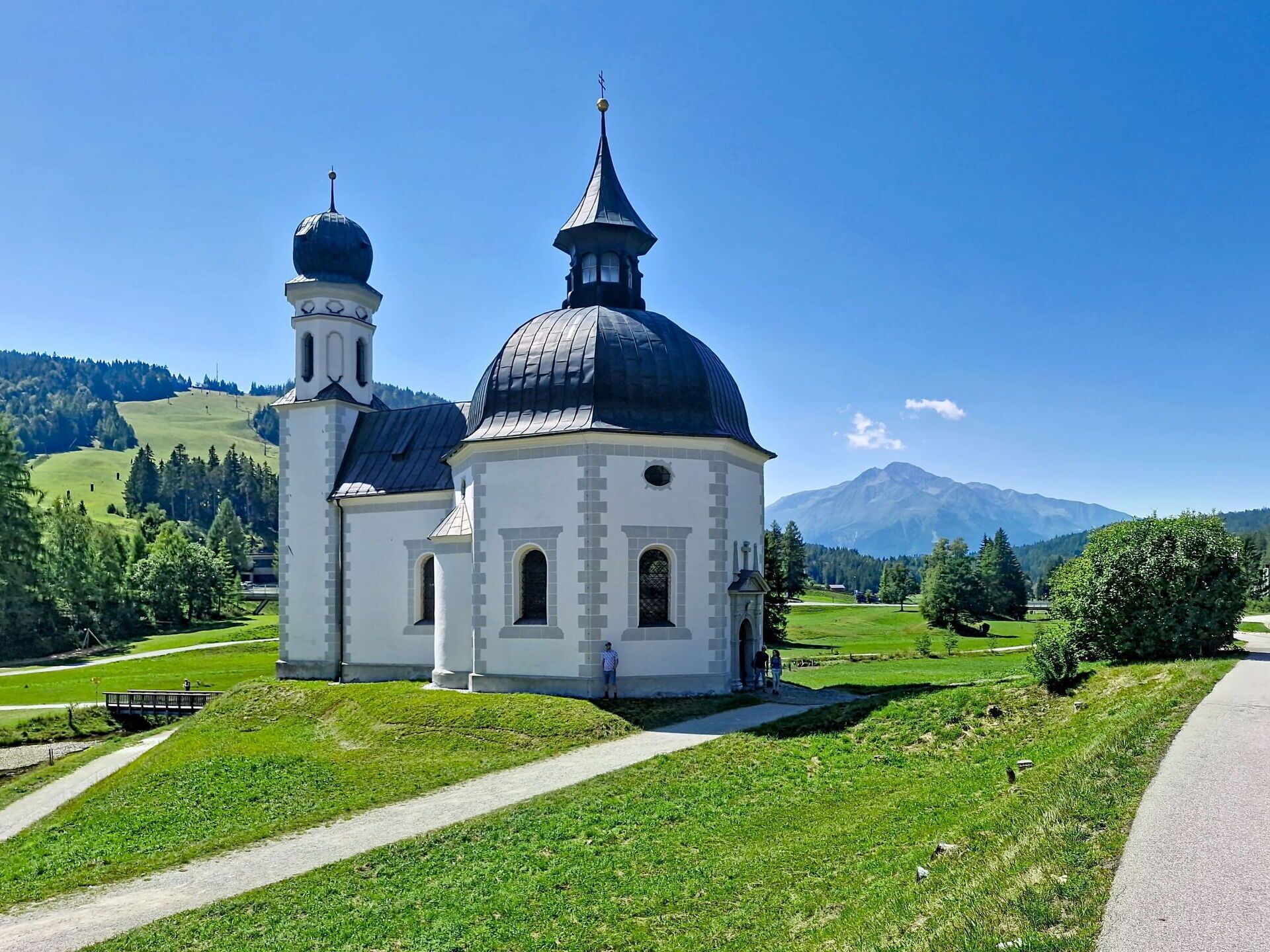 A white church with a large dome and two towers, surrounded by green hills and a clear sky.