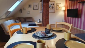 Een eetgedeelte met een houten tafel voor vier personen, omringd door stoelen. Achtergrond is een bank en een televisie.