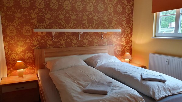 A double bed with white linens and two pillows, a wooden headboard, a bedside table with a lamp, and a window with a view of greenery outside. - Vacation apartment in the manor house - directly opposite the castle with terrace and WLAN (Gablenz)