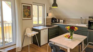 Une cuisine compacte avec un coin repas, une chaise blanche, une table sur laquelle se trouve un panier de fruits et un tableau accroché au mur.